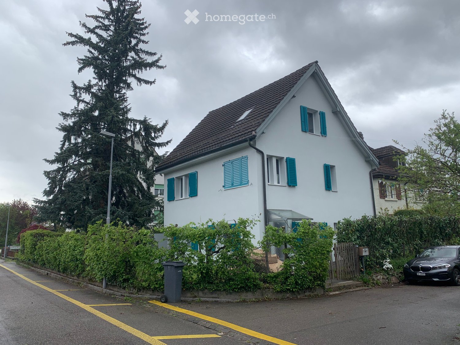House. Two-story house with white walls, blue shutters, and a brown tiled roof. The house is surrounded by greenery and has a parked car in front.