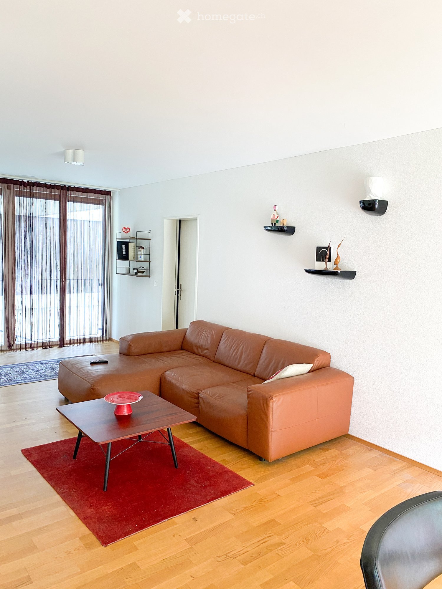 Living Room. Modern living room with a brown leather sofa, a wooden coffee table with a red plate, a small wooden table, wall-mounted shelves with decorative items, and a large window with curtains.