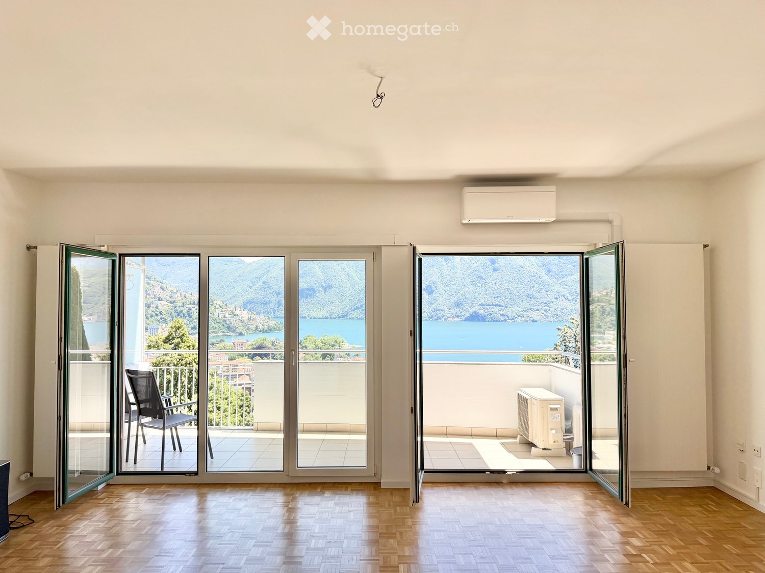 Empty room with wooden flooring, large sliding glass doors opening to a balcony with scenic lake and mountain views.