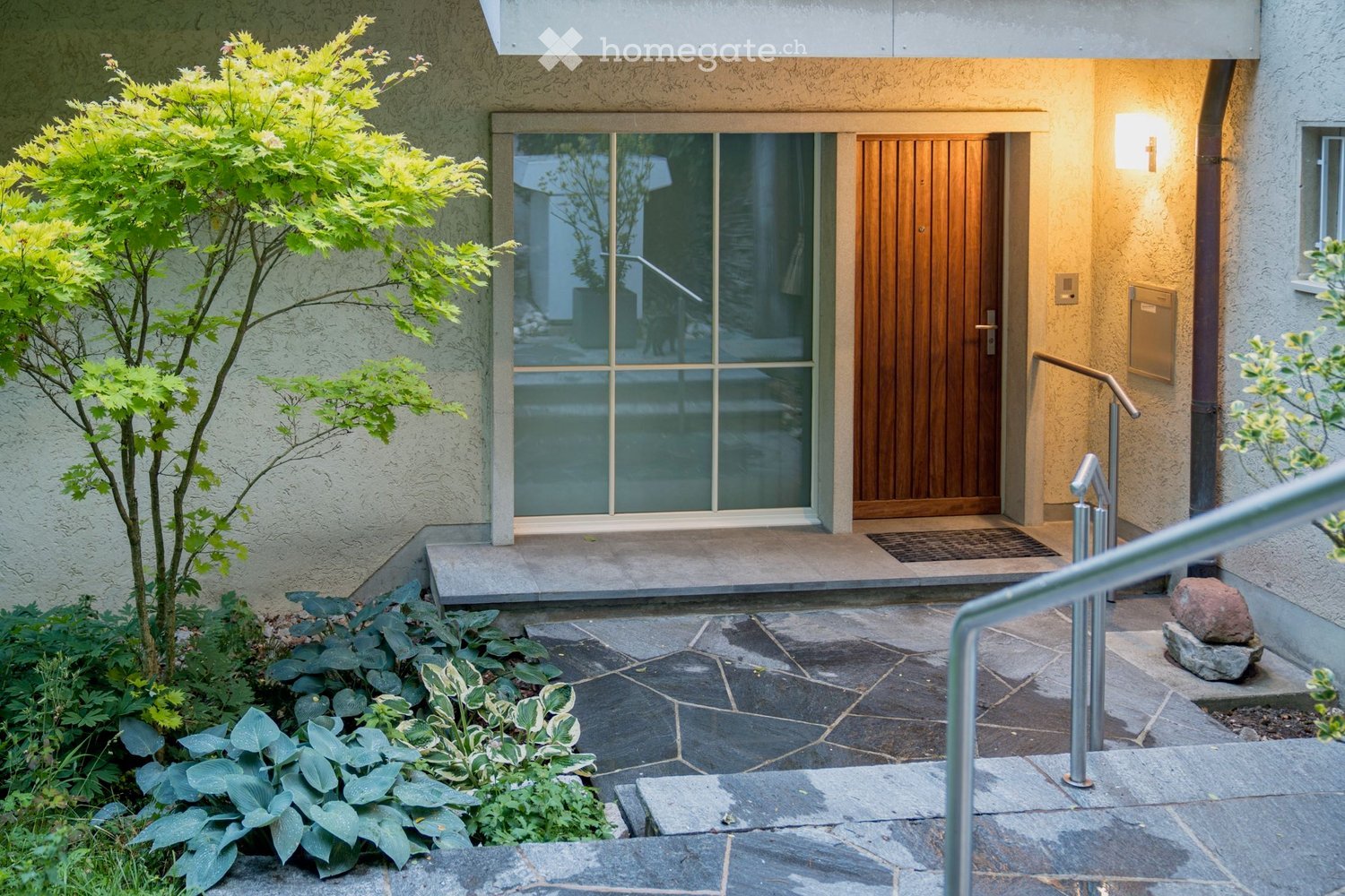 Garden / Outdoor Area. Entrance to a modern house with a raised ground floor, featuring a wooden door, glass panel, stone steps, and landscaped garden with shrubs and a small tree.