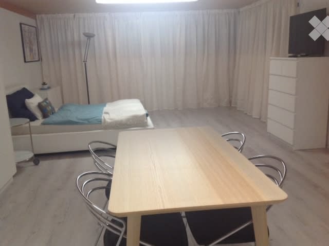 Furnished Room. A compact, well-lit room with a bed, a small table with chairs, a nightstand, a floor lamp, and a TV on a cabinet. The room features light-colored curtains and a neutral color palette.