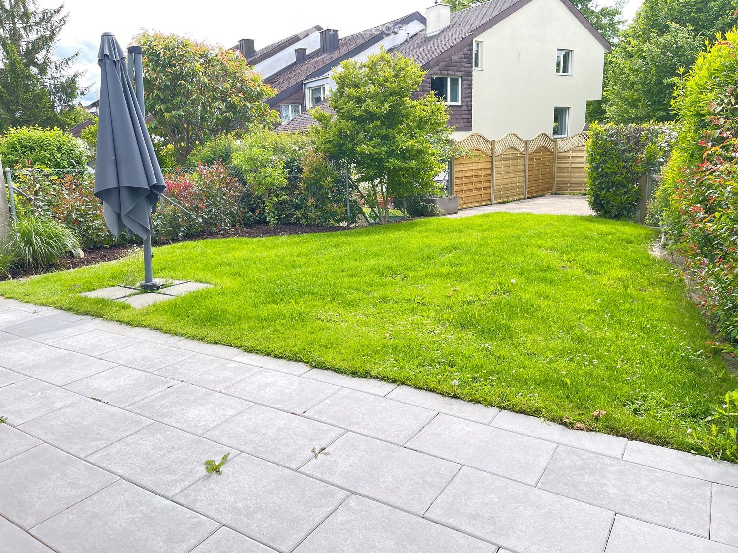 Well-maintained garden with a neatly trimmed lawn, surrounding trees, and a wooden fence. A closed umbrella is visible on the left side.
