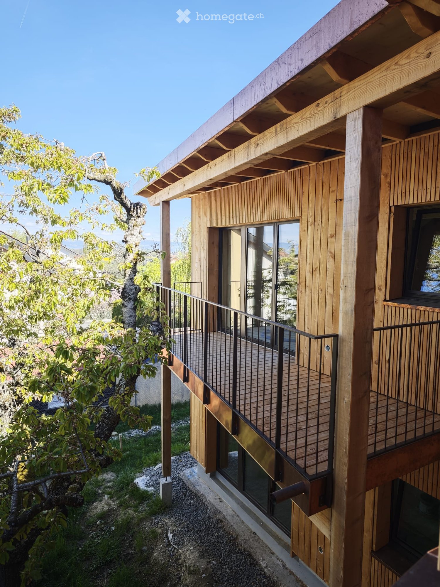 Building. Modern wooden house with a raised ground floor, large windows, and a balcony overlooking a garden area.