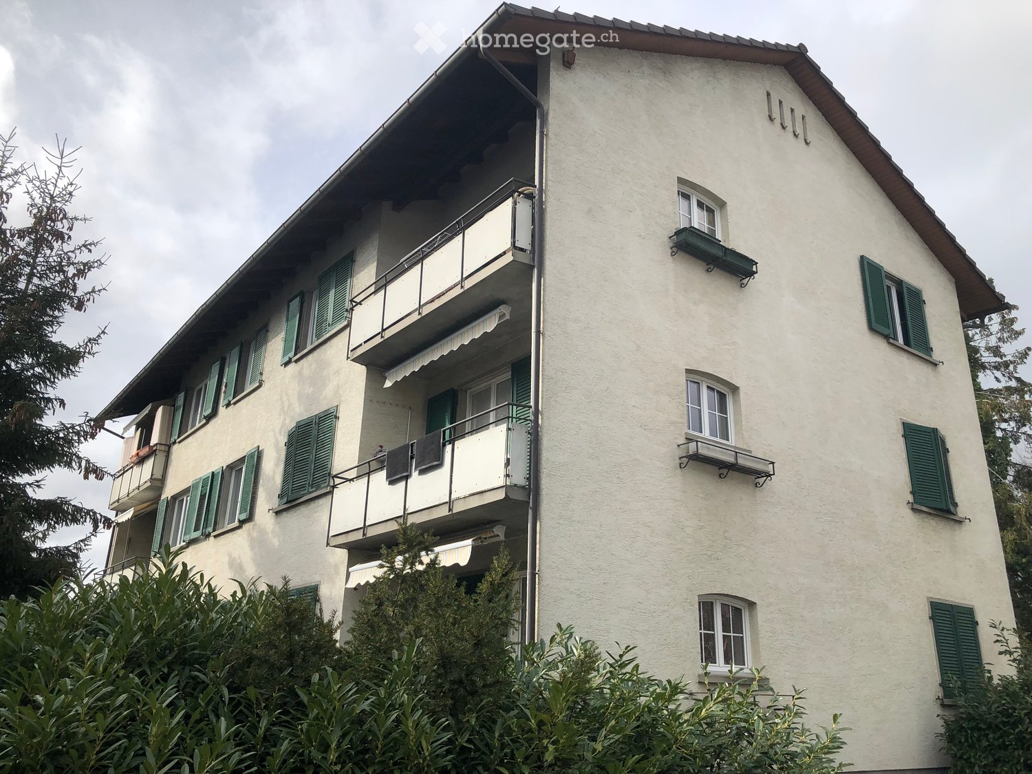 Building. A multi-story residential building with light-colored walls, green shutters, and multiple balconies. The building has a classic architectural style with a pitched roof and visible laundry hanging on one of the balconies.