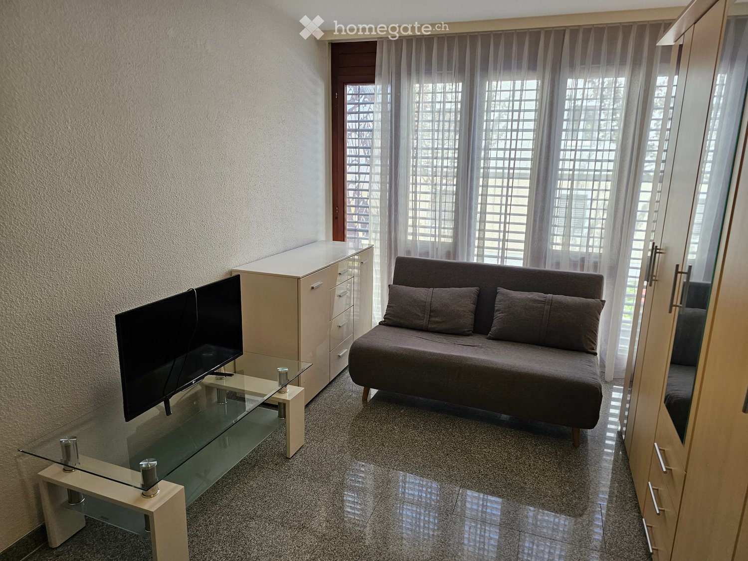 Gray sofa, flatscreen TV, glass table, wooden cabinets, windows with white curtains