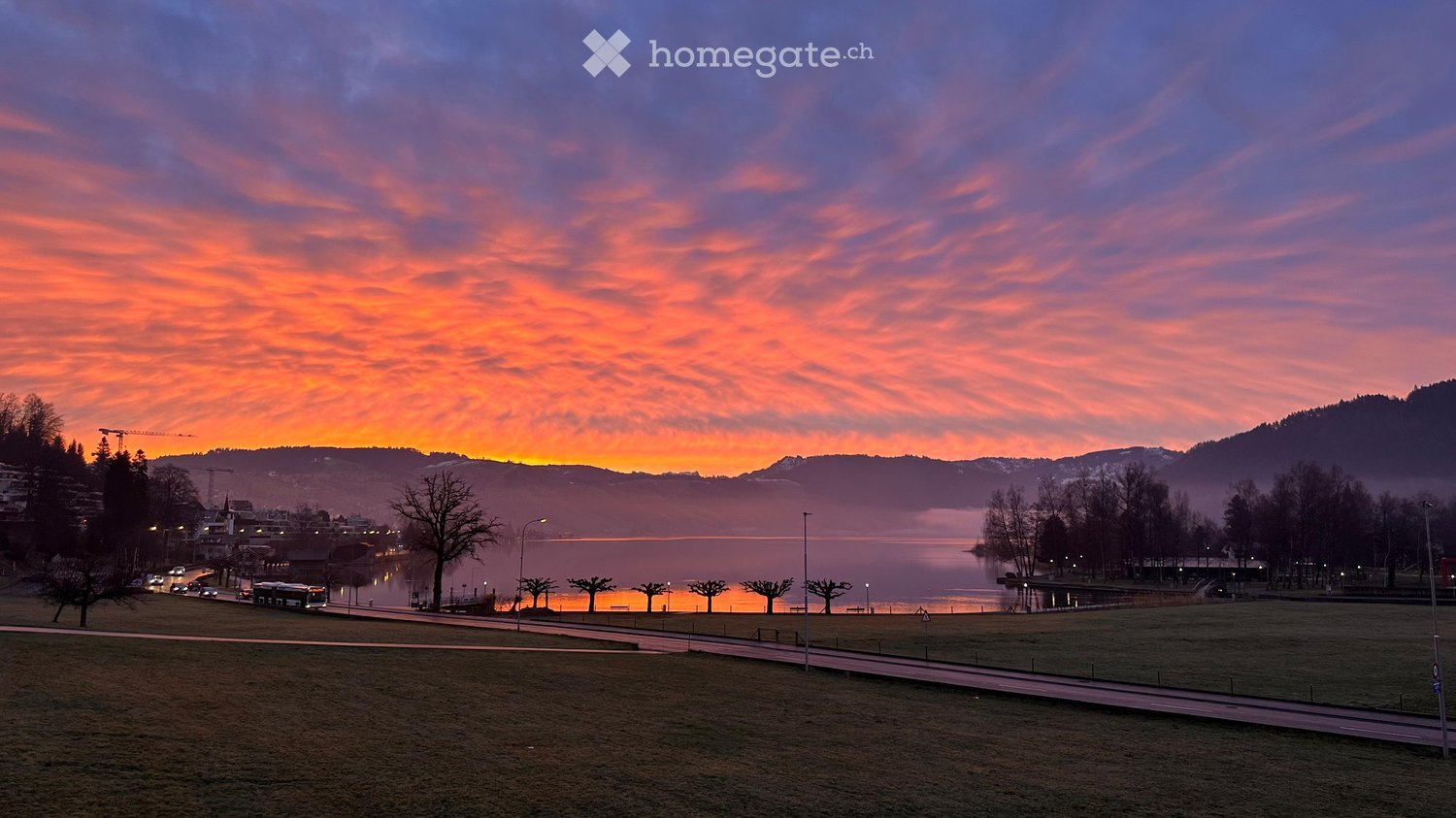 Garden / Outdoor Area. A serene lakeside scene at sunset, with calm water reflecting the vibrant orange and pink sky, surrounded by trees and distant hills.
