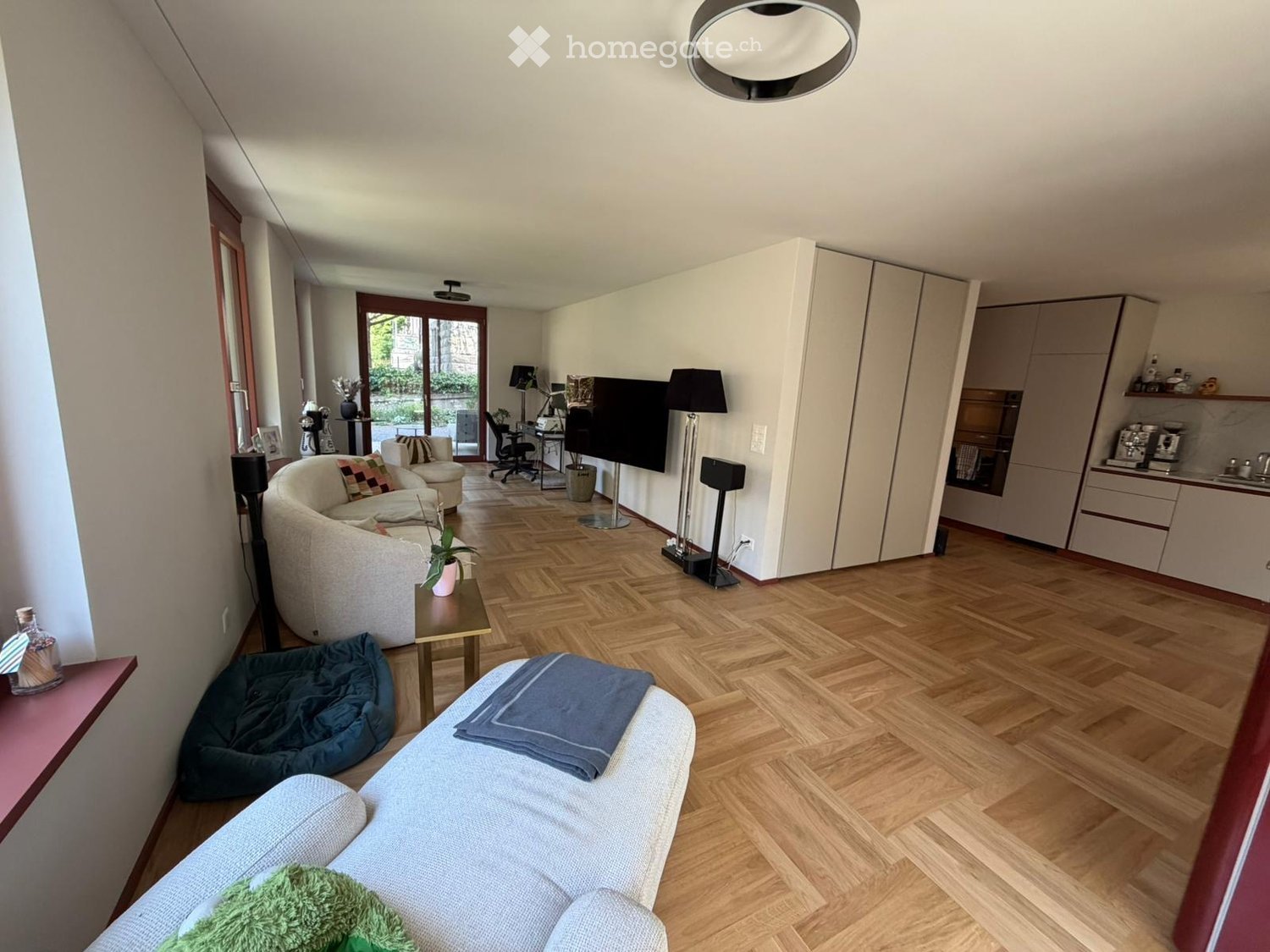 Furnished Room. A modern, well-lit living room with a large sofa, a chair, a coffee table, a TV, and a small kitchen area. The room features wooden flooring, large windows, and a cozy atmosphere.
