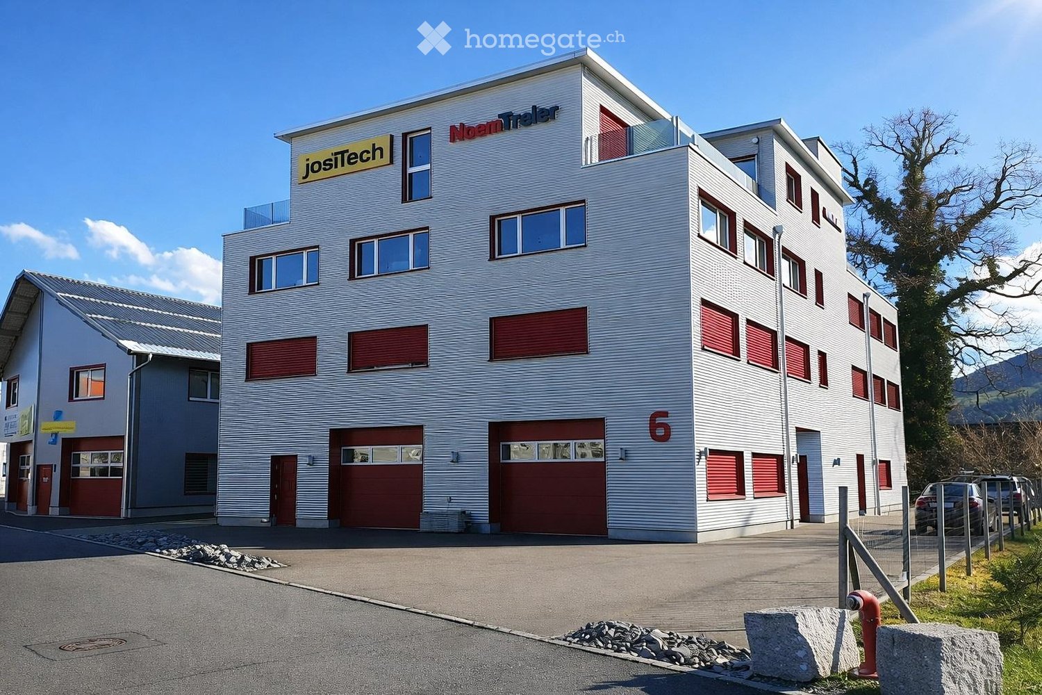 Building. Modern multi-story building with white exterior, red garage doors, and signage for 'JosTech' and 'NoamTeler'. Features large windows, balconies, and a parking area.