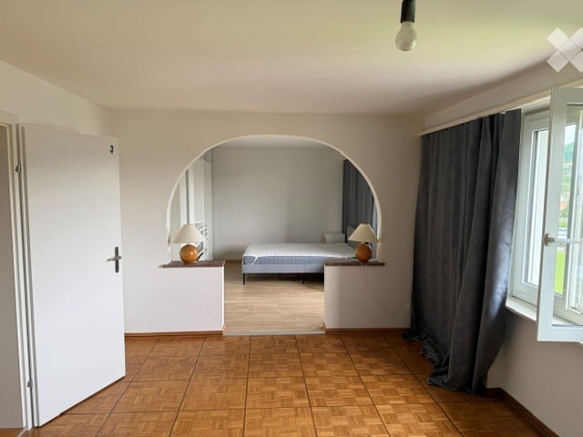 Furnished Room. A furnished room with a bed, nightstands, and lamps. The room features wooden flooring and large windows with curtains.
