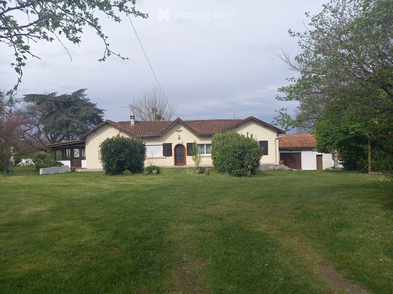 House. Single-story house with a tiled roof, beige walls, and a garage. The house is surrounded by a well-maintained lawn and trees.