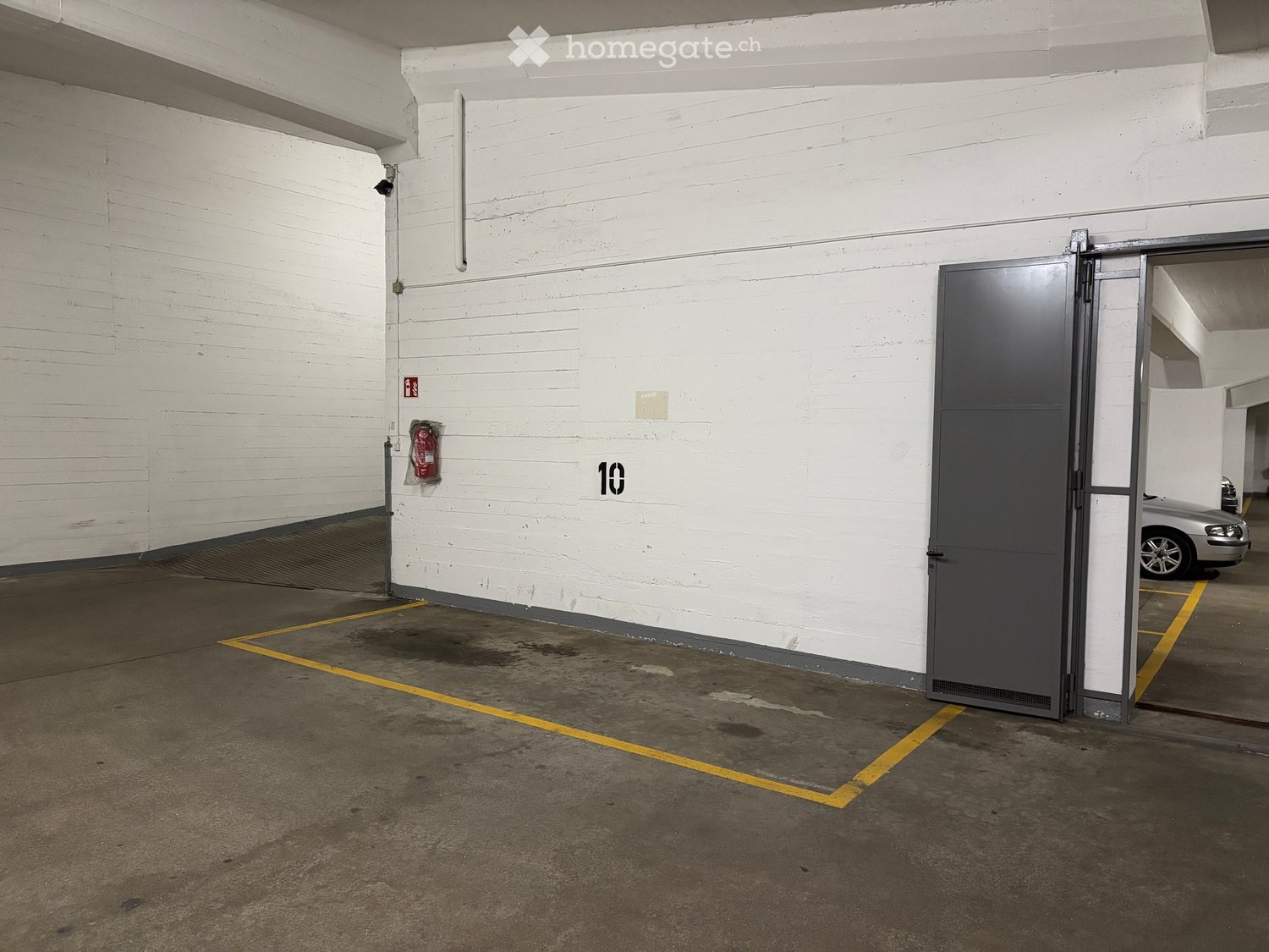 Other. Empty parking space in a garage with white brick walls, a large grey door, and a silver car partially visible in the background.