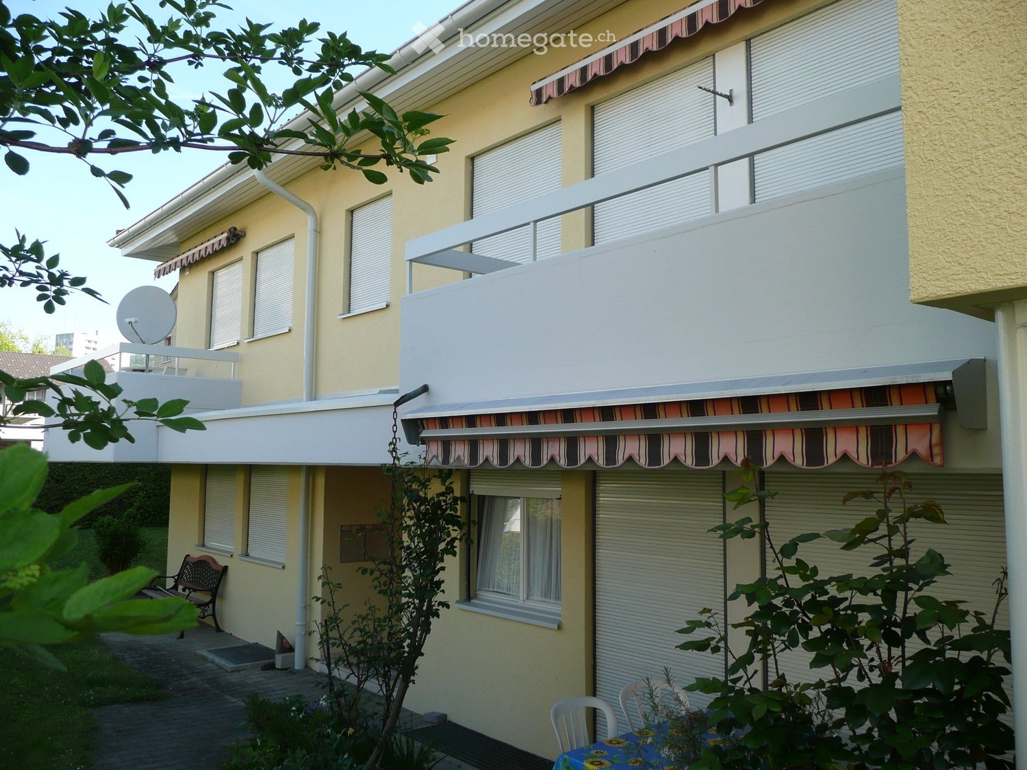 Building. Two-story residential building with balconies, striped awnings, and satellite dish. Lower level has a small patio with a table and chairs.