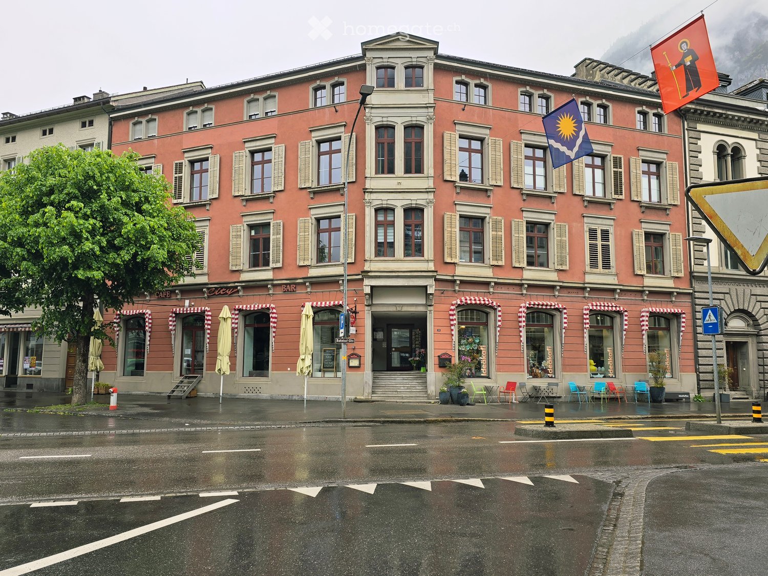 Building. A multi-story building with a pink facade, multiple windows, and several storefronts including a cafe and shops. Flags are displayed on the building, and outdoor seating is visible in front of the shops.