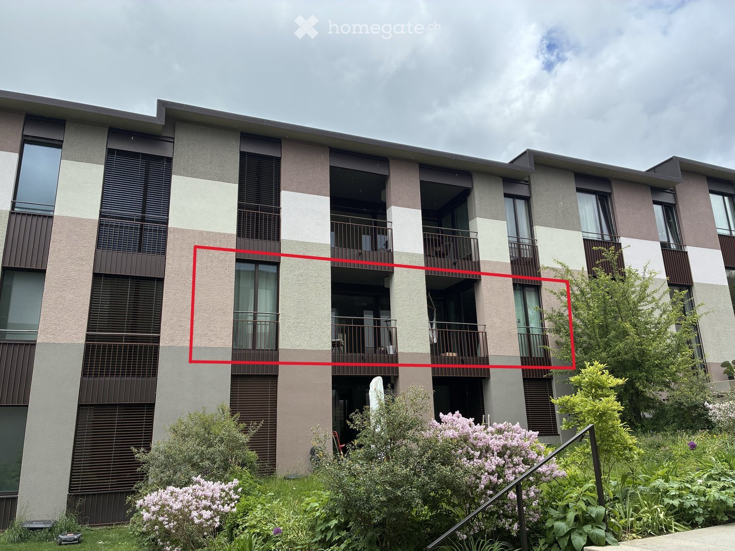 Building. Modern multi-story residential building with balconies, large windows, and contemporary design. Features include a mix of beige and brown tones, vertical blinds, and a landscaped garden area with flowering shrubs and trees.