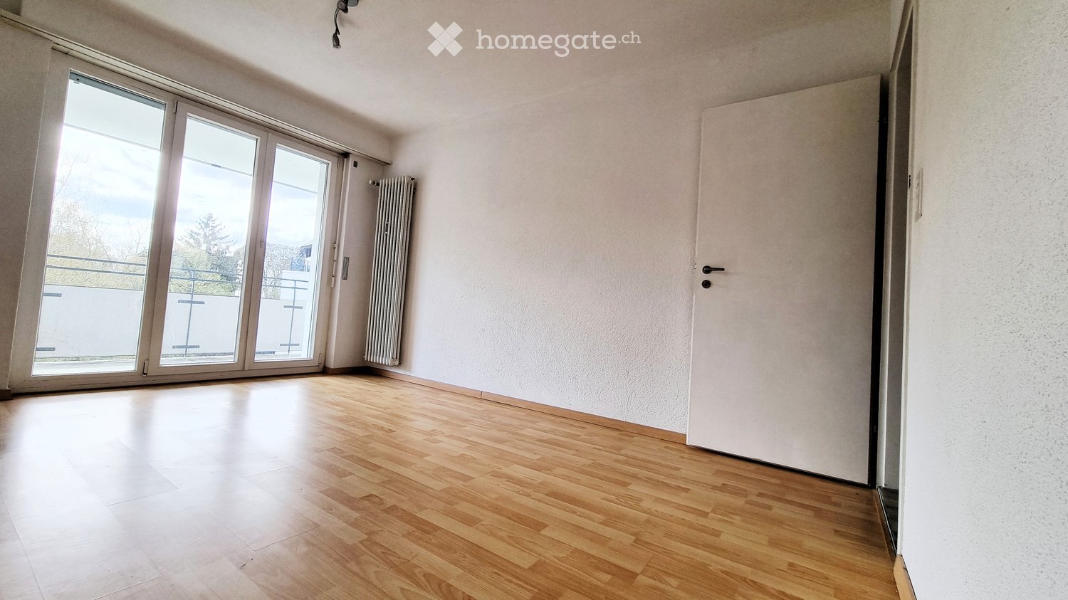 Unfurnished Room. Empty room with wooden flooring, large sliding glass doors leading to a balcony, white walls, and a radiator.
