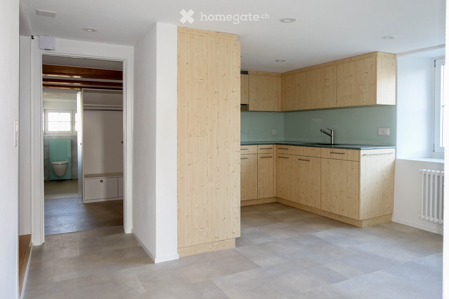 Furnished Room. Modern kitchen with light wooden cabinets, a glass backsplash, and a stainless steel sink. Adjacent bathroom with a toilet and a window.