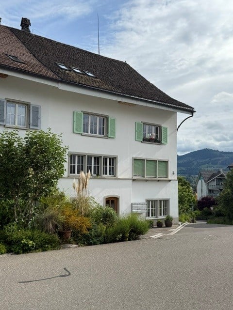 House. A three-story white house with green shutters, multiple windows, and a brown tiled roof. The house is surrounded by greenery and has a driveway in front.