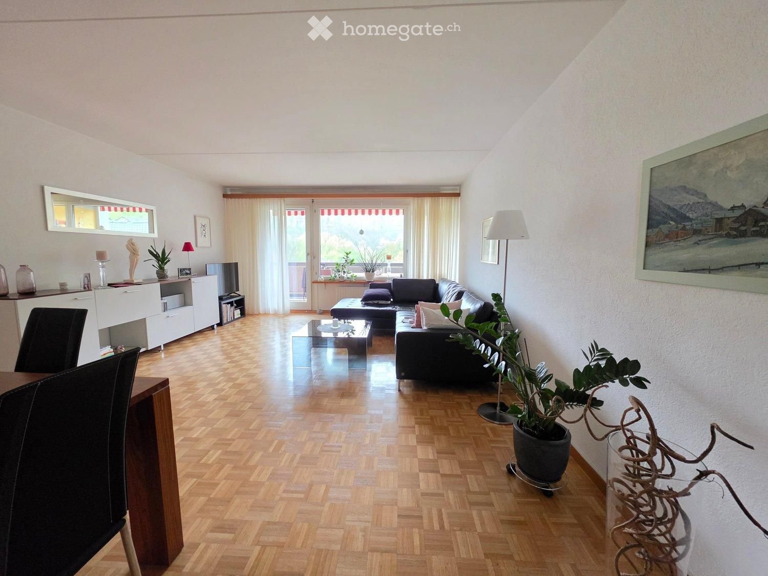 Living Room. Bright living room with wooden flooring, large windows, a black sectional sofa, coffee table, dining chairs, and decorative plants.