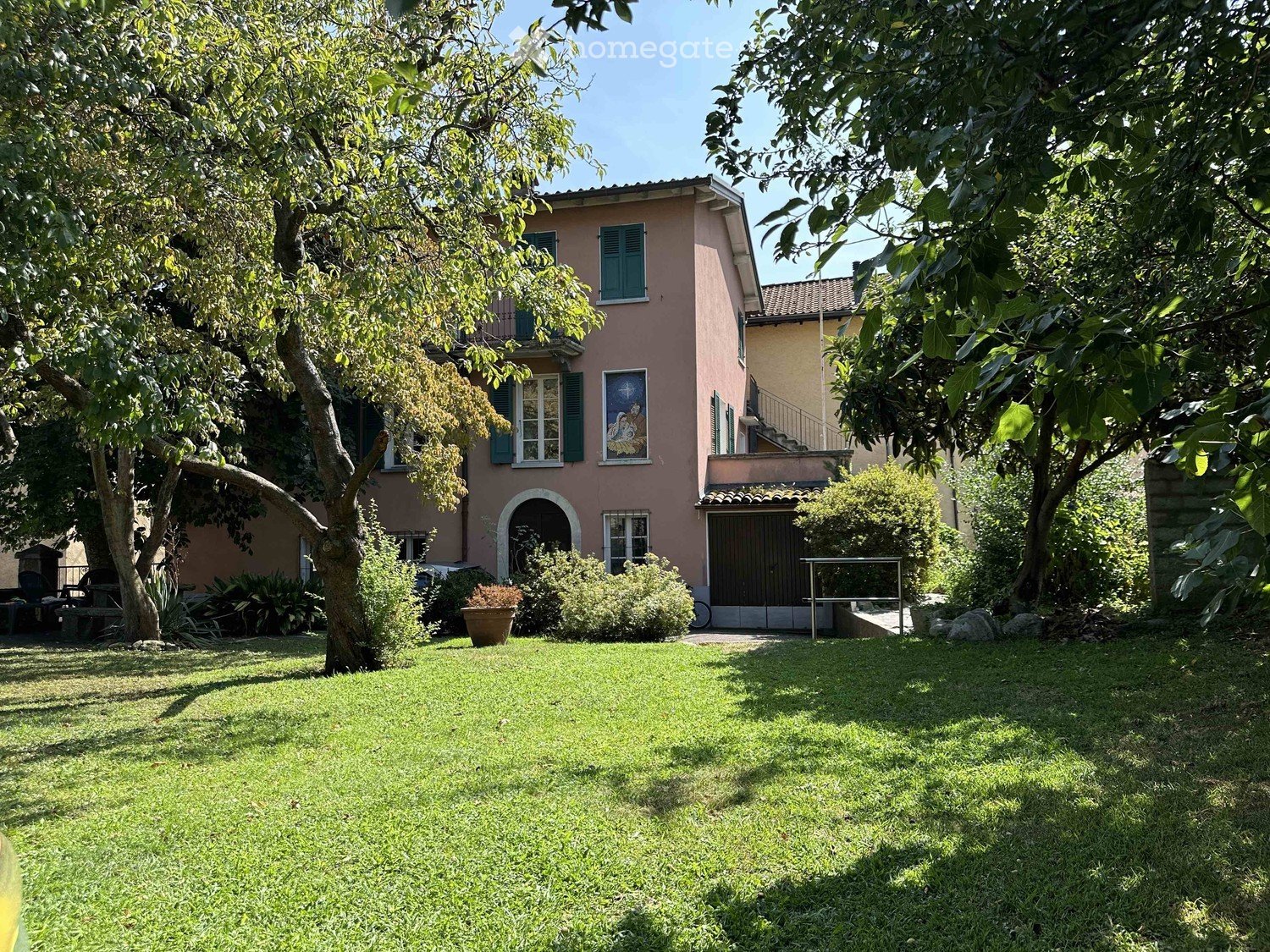 Lush garden with a well-maintained lawn, trees, shrubs, and a pink house with green shutters in the background.