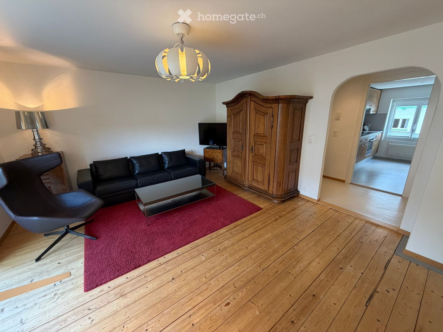 Living room with wooden floor, black couch, glass coffee table, wooden cabinet, lamp, TV, wooden chair, red carpet