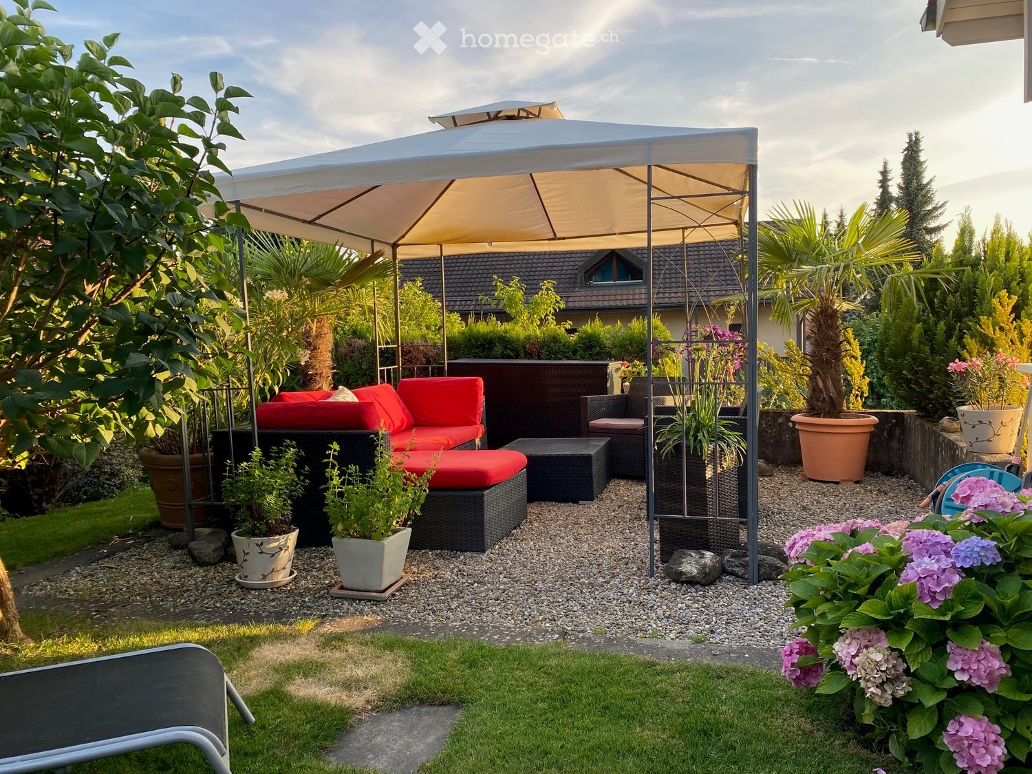 Garden / Outdoor Area. A well-maintained garden with a red outdoor sofa set, a black table, a white umbrella, and various potted plants and flowers.