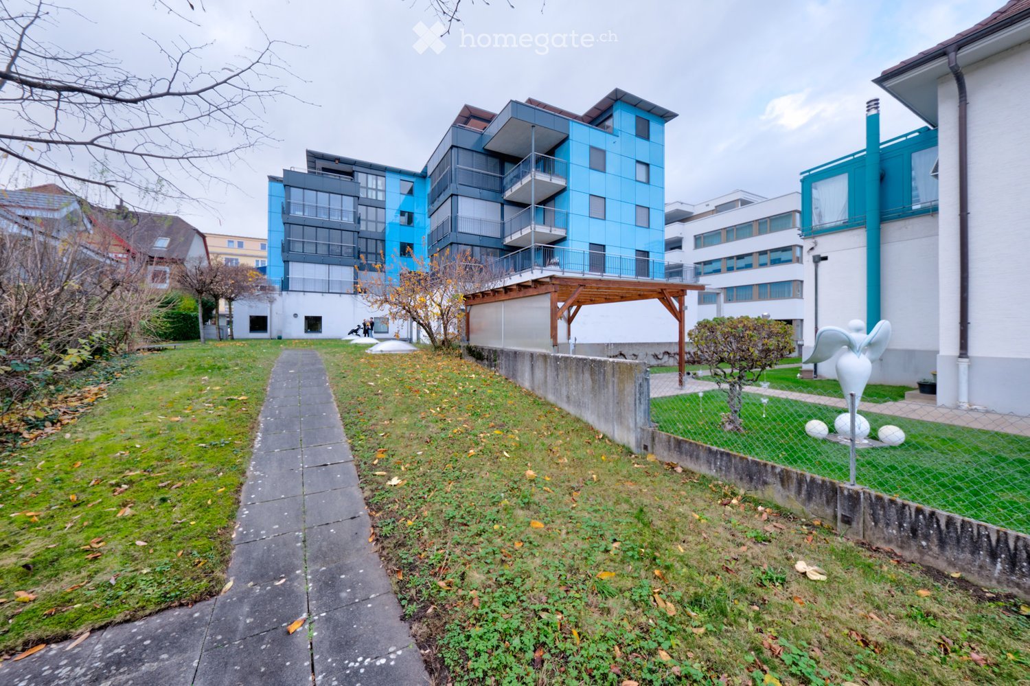 Modern apartment building with blue facade, multiple balconies, and a small garden area.