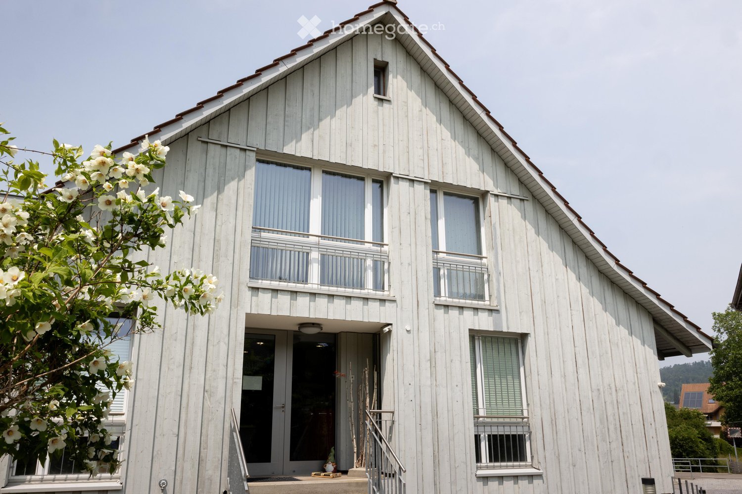 House. Two-story white wooden house with a steeply pitched roof, multiple windows, and a small balcony on the upper floor. The entrance features a glass door and a small staircase leading up to it. A flowering tree is visible on the left side.