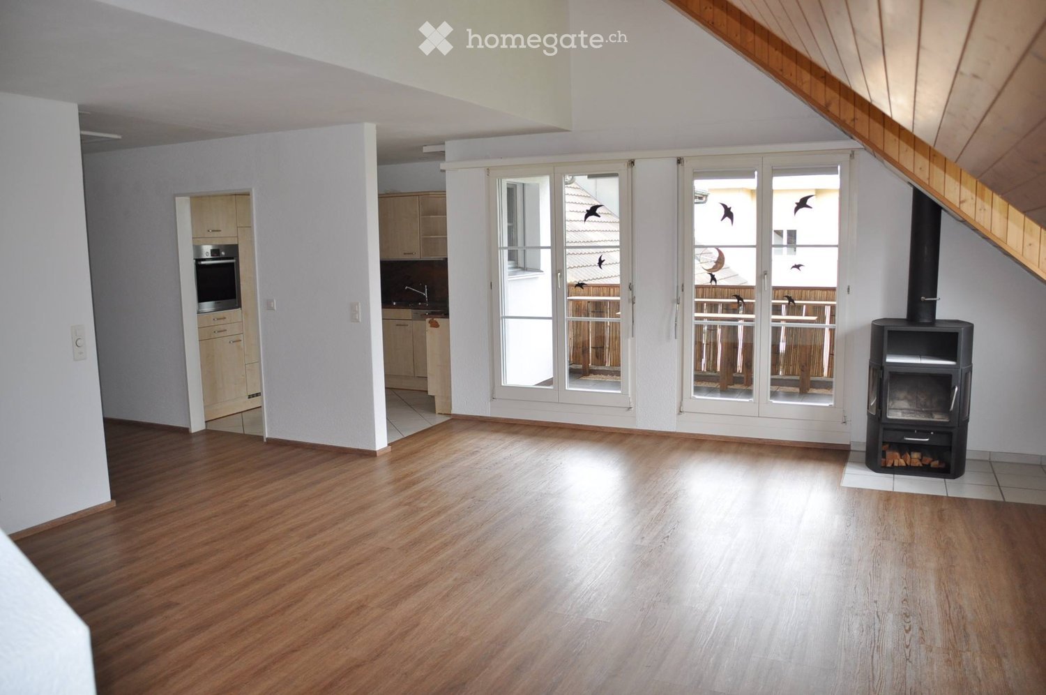 Furnished Room. Bright living space with wooden flooring, large glass doors leading to a balcony, fireplace, and an adjacent kitchen area with cabinets and an oven.