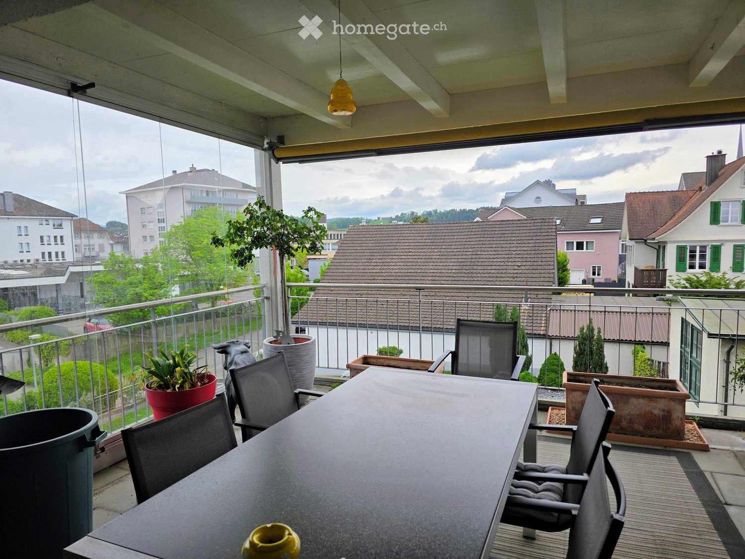 Balcony / Terrace. Balcony with a table, chairs, and potted plants, overlooking a residential area with houses and greenery.