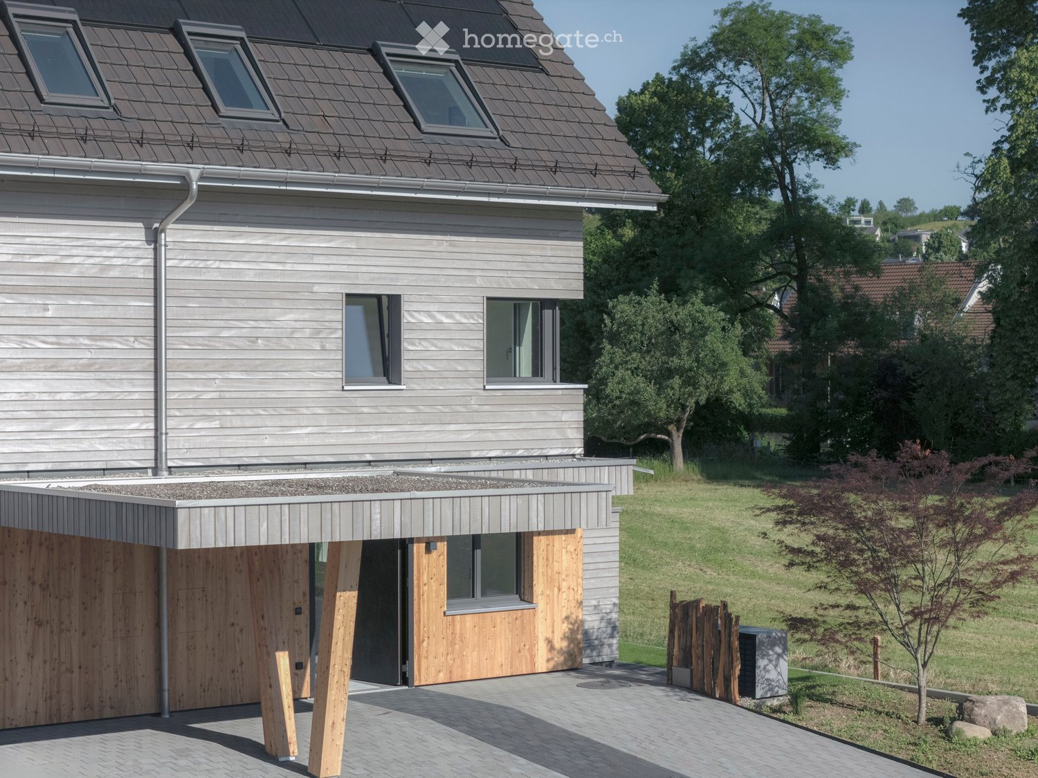 Modern two-story house with grey wooden siding, large windows, and a flat roof terrace. Features a covered entrance with wooden pillars and a paved driveway.