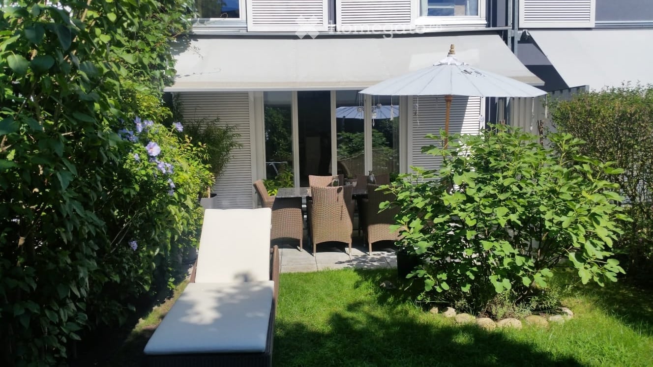 2-story house with white exterior, large windows, patio with rattan furniture and umbrella, surrounded by lush greenery and a small lawn