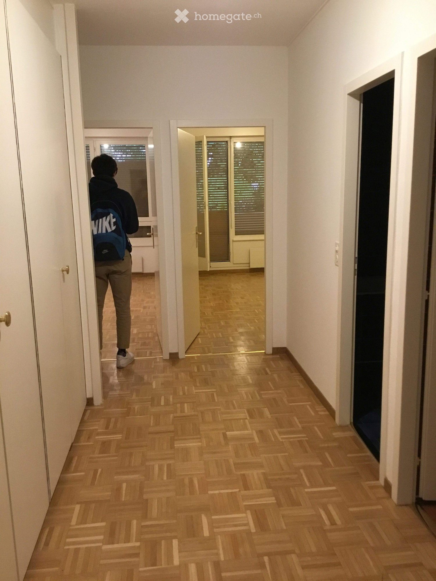 Corridor / Hallway. A long, narrow hallway with wooden flooring and white walls, leading to two open doors that reveal rooms with large windows and blinds.