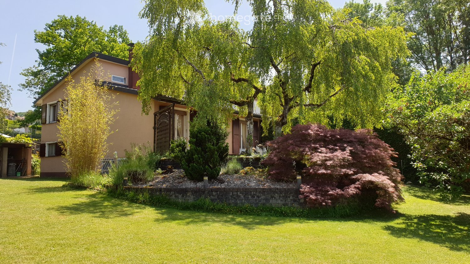 Single-story house with beige exterior, surrounded by a well-maintained garden with trees, shrubs, and a lawn.