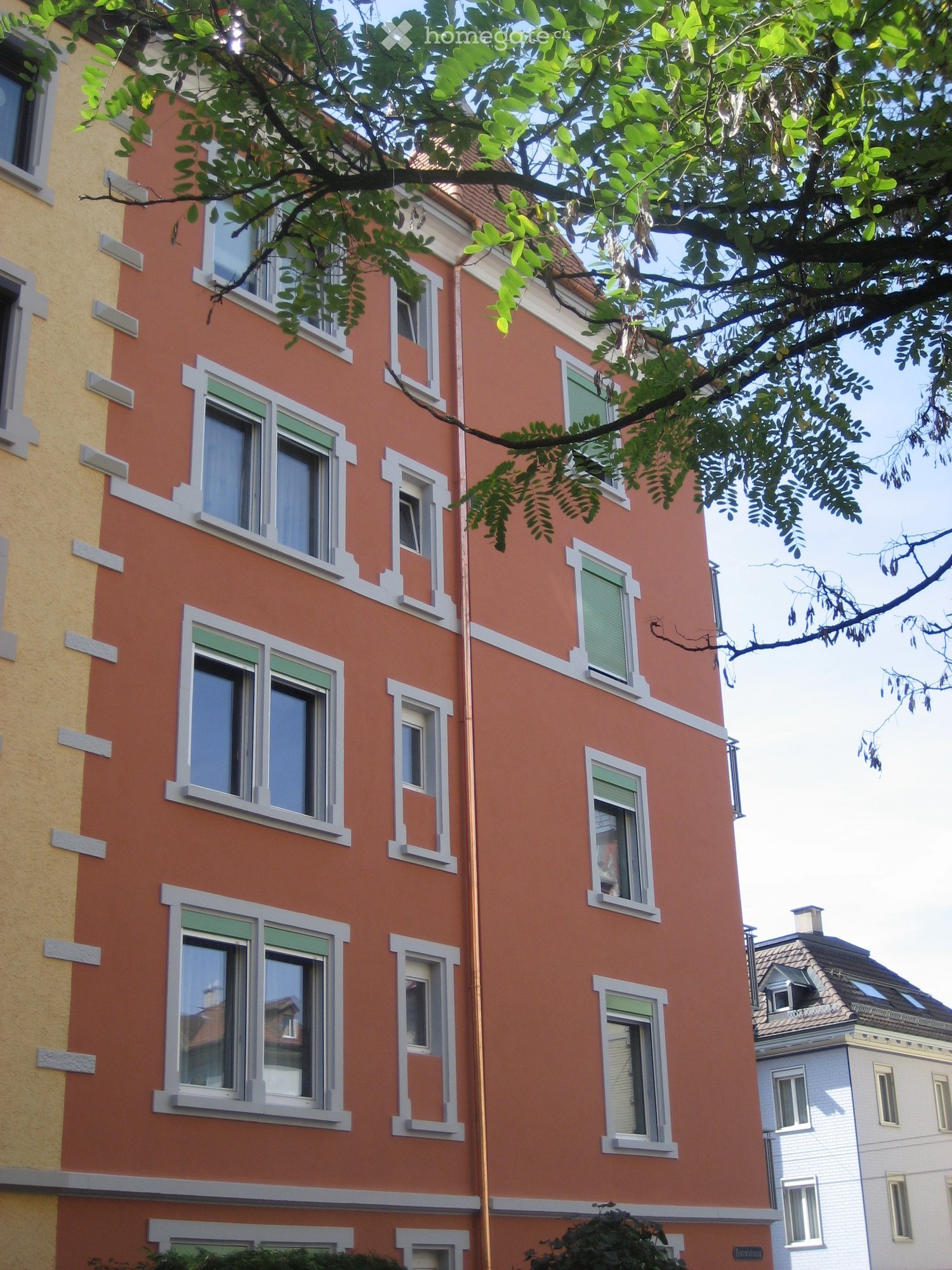 Building. A multi-story residential building with orange and yellow facades, multiple windows with white frames, and green shutters. The building is partially shaded by tree branches.