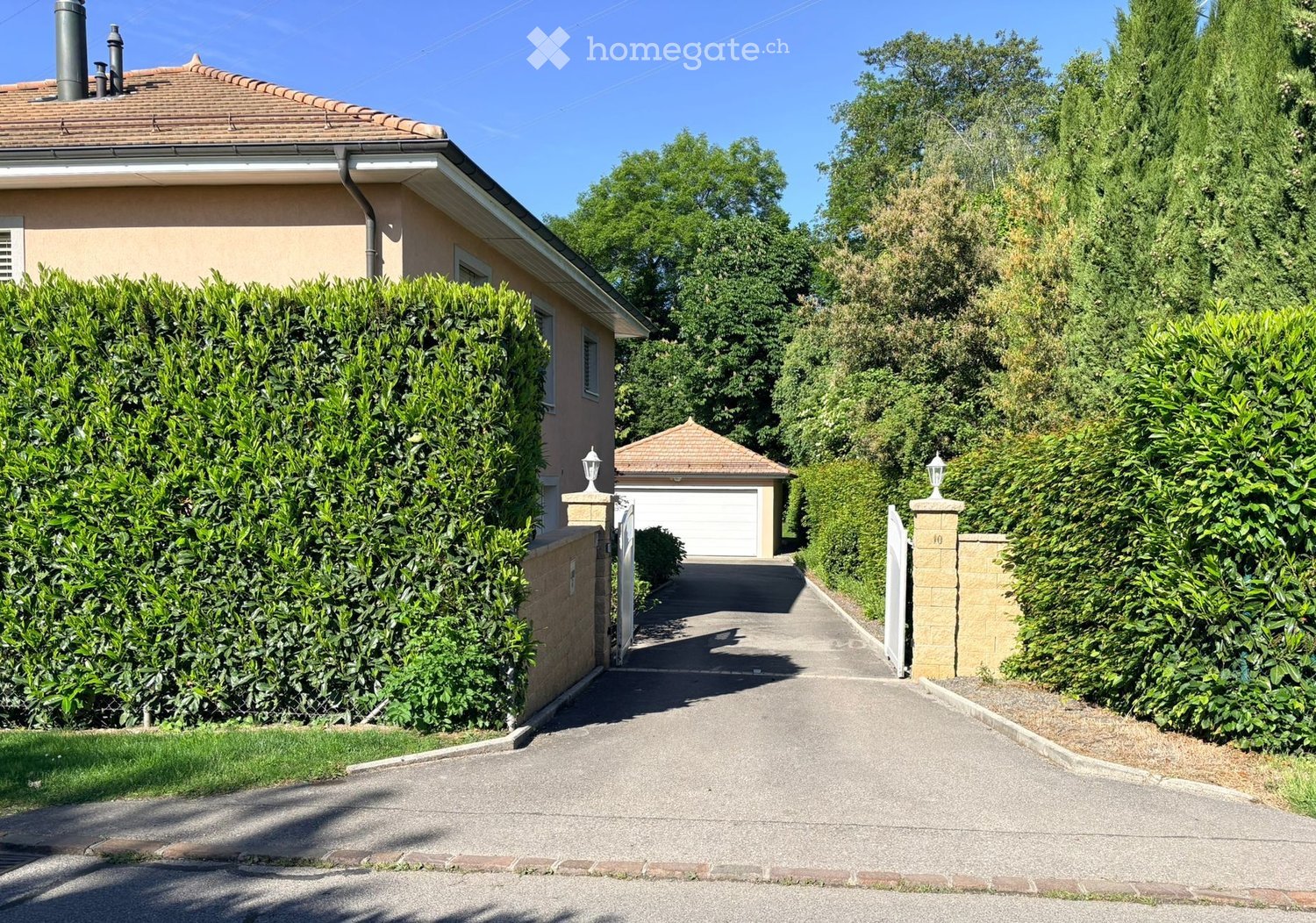House. A two-story house with a tiled roof, beige exterior, and a well-maintained hedge-lined driveway leading to a garage.