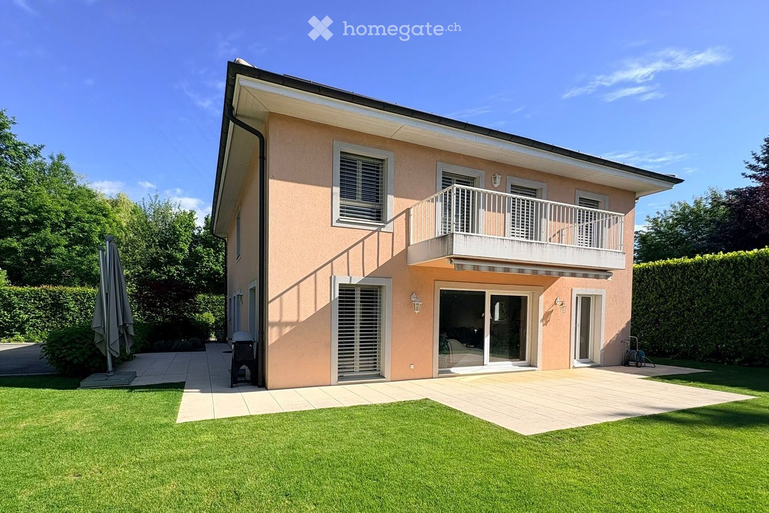 House. Two-story house with light pink exterior, white-framed windows, and a balcony on the upper level. Features a well-maintained lawn, patio area, and outdoor furniture.