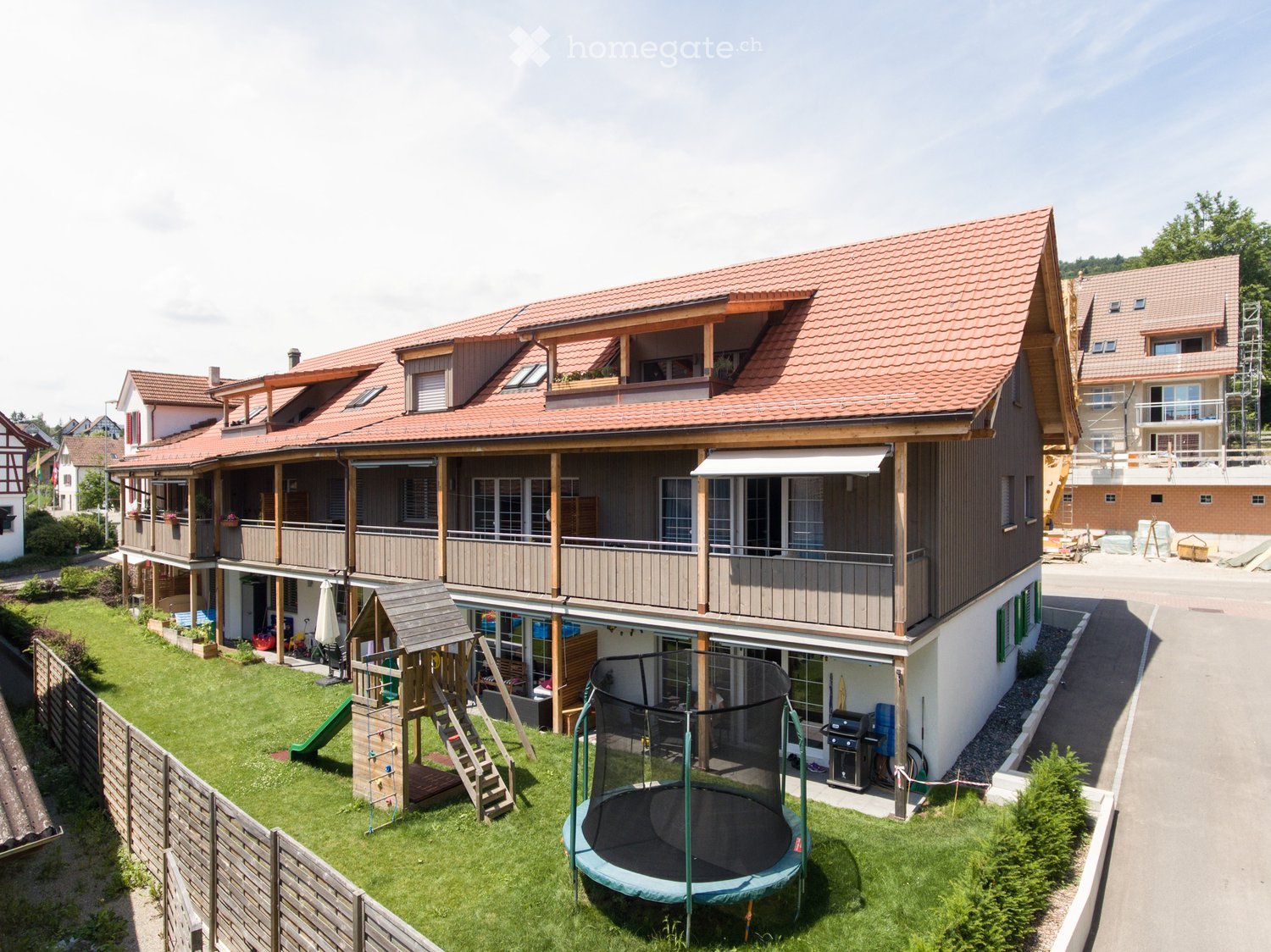 House. Two-story house with red-tiled roof, balconies, and a fenced backyard featuring a trampoline, playground equipment, and a barbecue area.