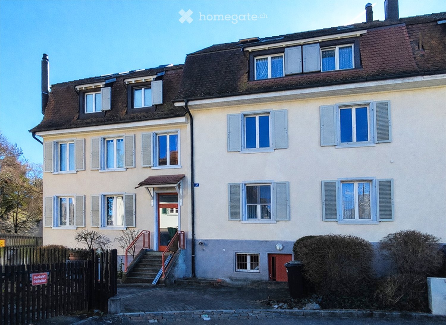 3 story building, beige walls, brown roof, multiple windows, stairs leading to the entrance, small garden in front