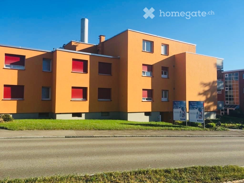 Building. Modern orange-colored building with multiple windows and a flat roof, surrounded by a grassy area and signage.