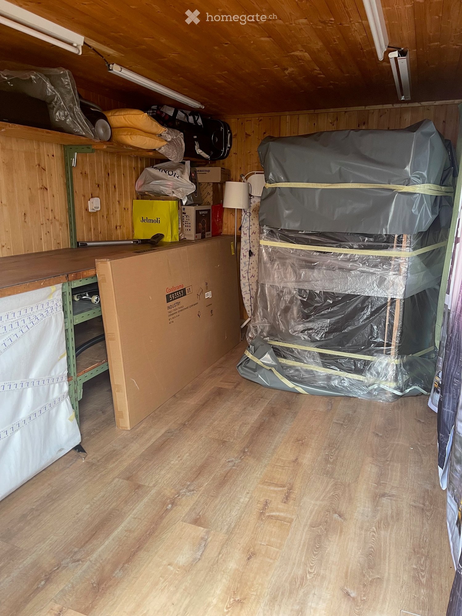 Unfurnished Room. An attic or storage room with wooden walls and flooring, featuring shelves and various stored items including boxes, a covered appliance, and a mattress wrapped in plastic.