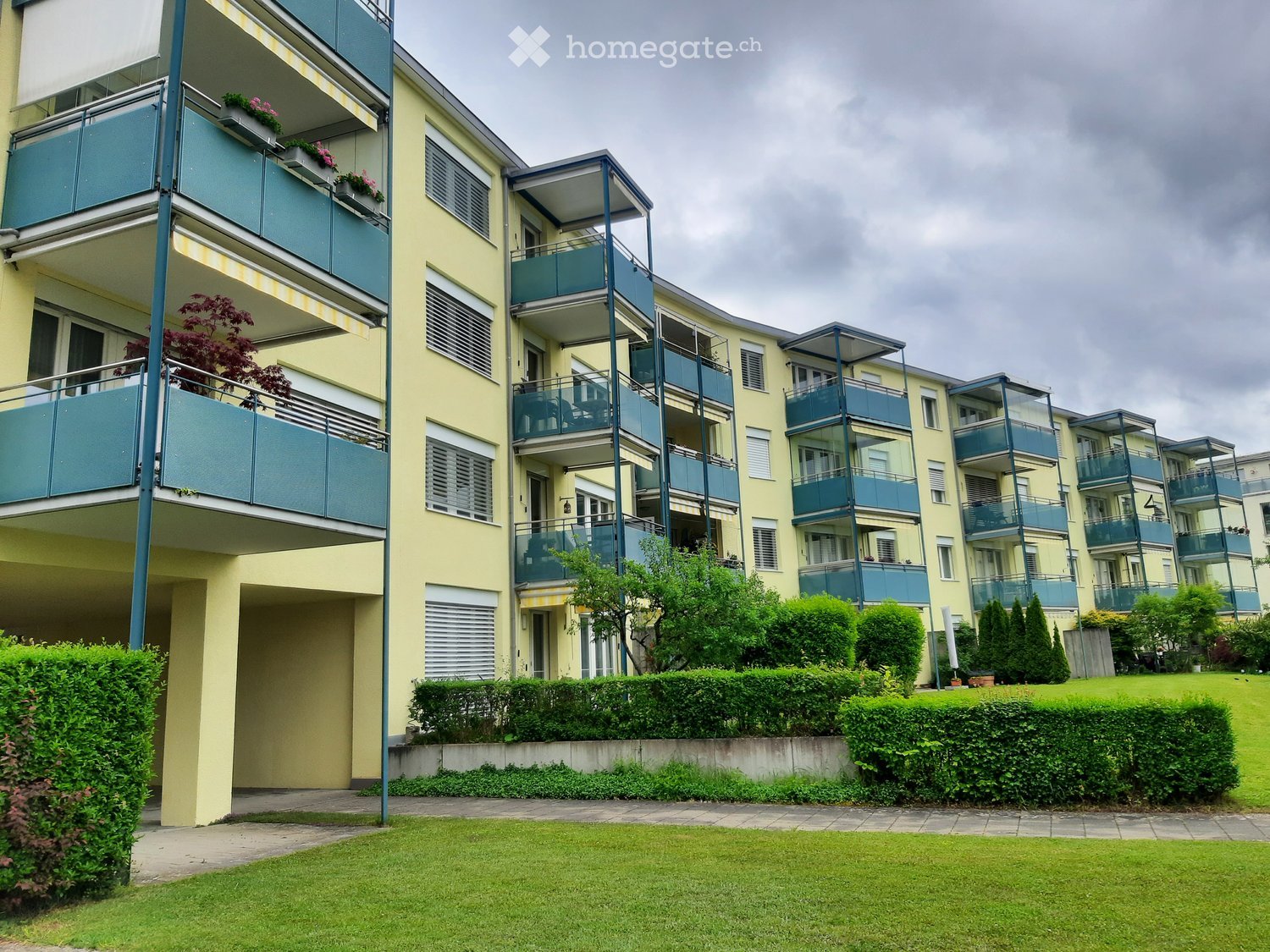 Building. Multi-story residential building with yellow facade, blue balconies, and surrounding greenery.