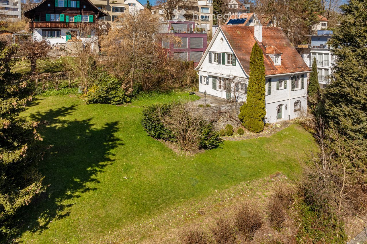 Garden / Outdoor Area. Aerial view of a well-maintained garden with a large green lawn, surrounded by trees and bushes. Several houses with distinct architectural styles are visible in the background, including a white house with green shutters and a red-