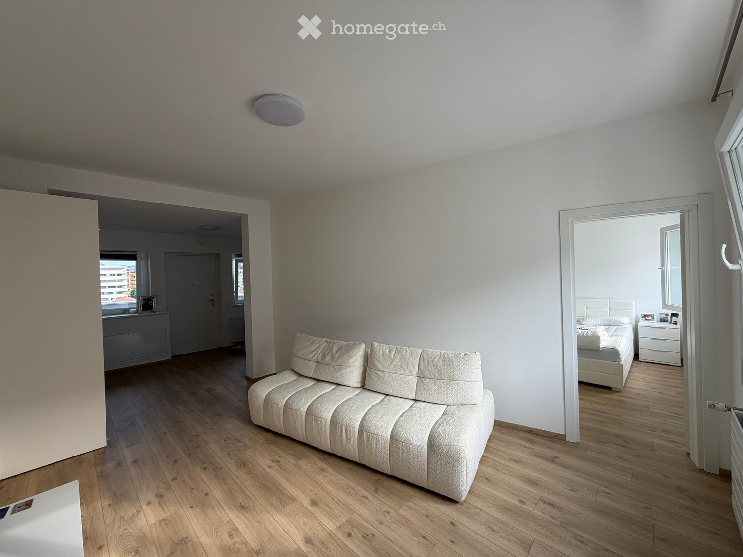 Furnished Room. Modern living room with a white sofa, wooden flooring, and an open doorway leading to a bedroom with a bed and nightstand.