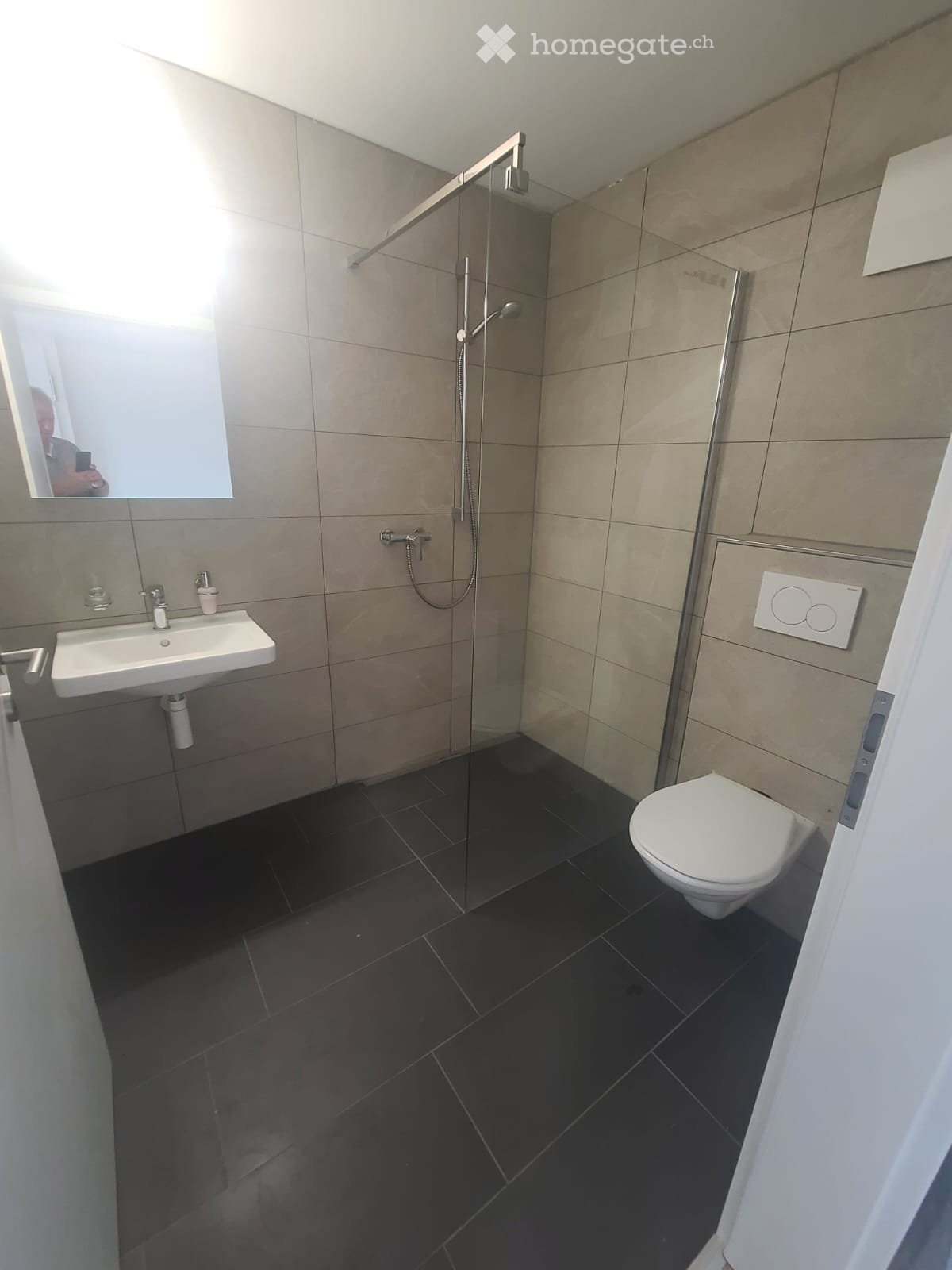 Bathroom. Modern bathroom with a glass-enclosed shower, a white sink, a toilet, and a large mirror. The walls are tiled in a neutral color, and the floor is dark tiled.