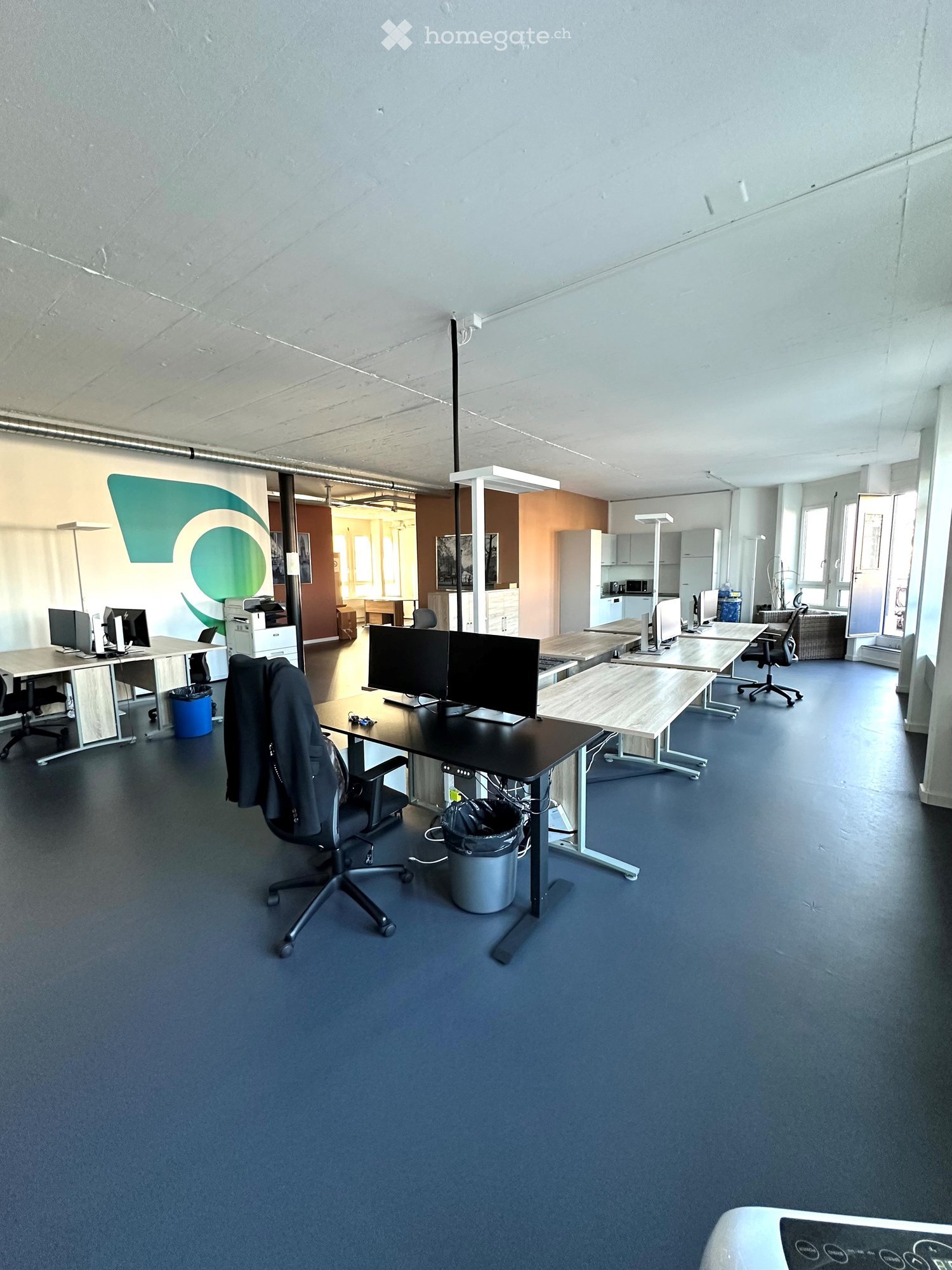 Furnished Room. Modern office space with desks, chairs, computer monitors, and a printer. The room features a blue carpeted floor and white walls.