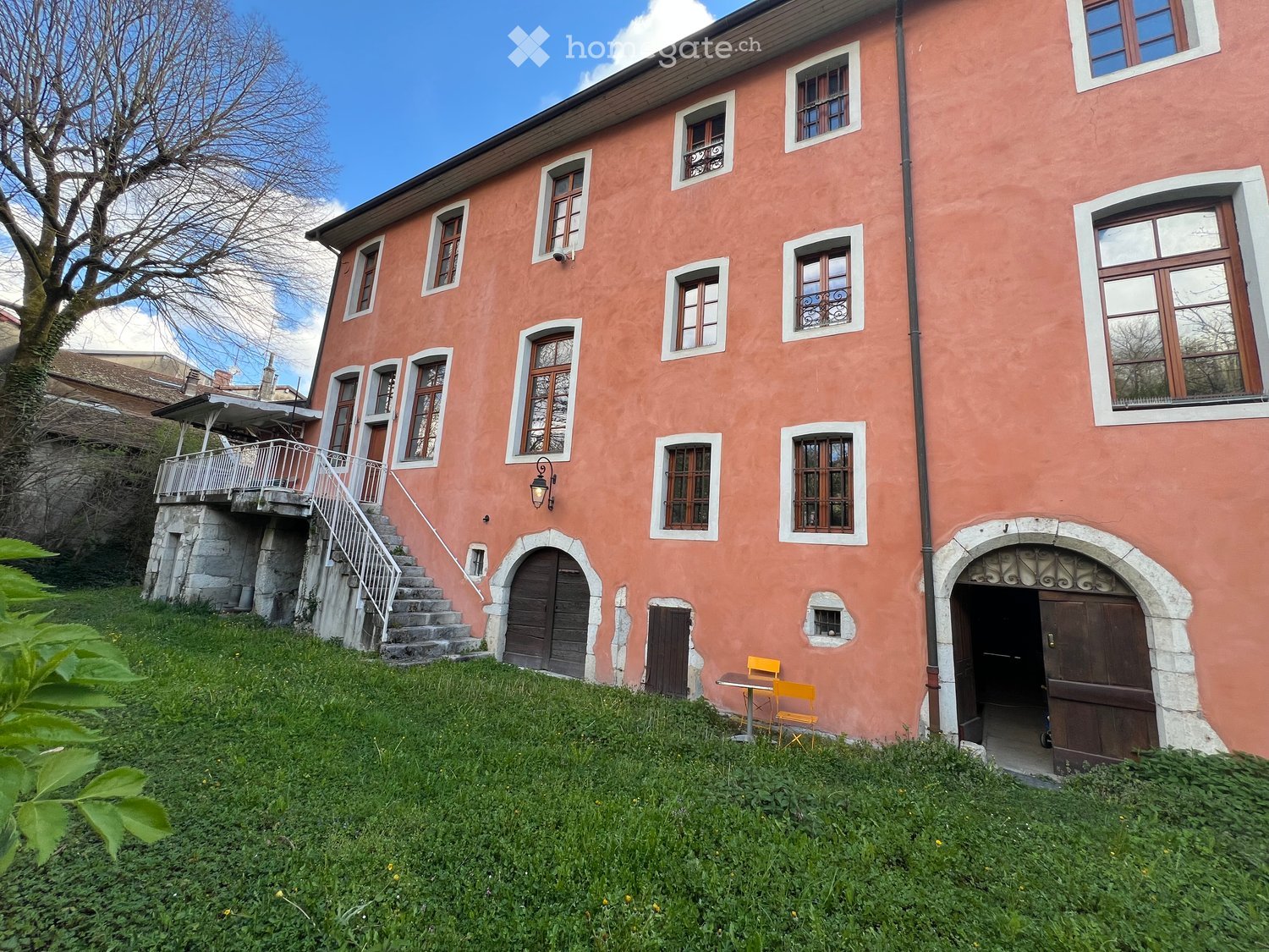 Building. A two-story pink building with multiple windows, a raised ground floor, and a small outdoor seating area with a table and chairs.