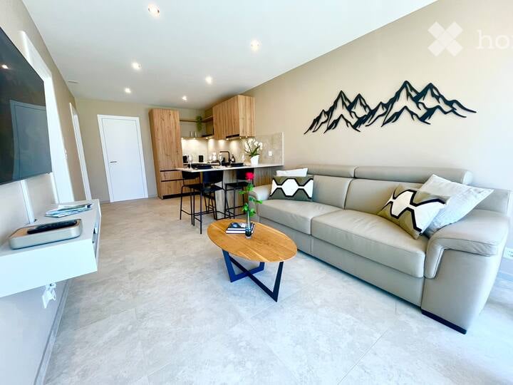 Furnished Room. Modern open-plan living and dining area with a light grey sofa, wooden coffee table, flat-screen TV, and a small kitchen area with bar stools and wooden cabinets.