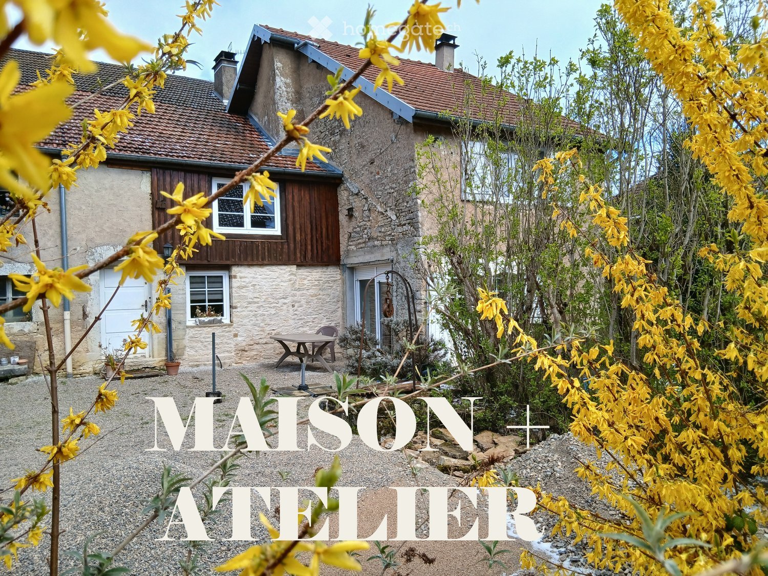 Garden / Outdoor Area. A rustic stone house with a red-tiled roof, surrounded by a garden with yellow flowering shrubs and a small patio area with a table and chairs.