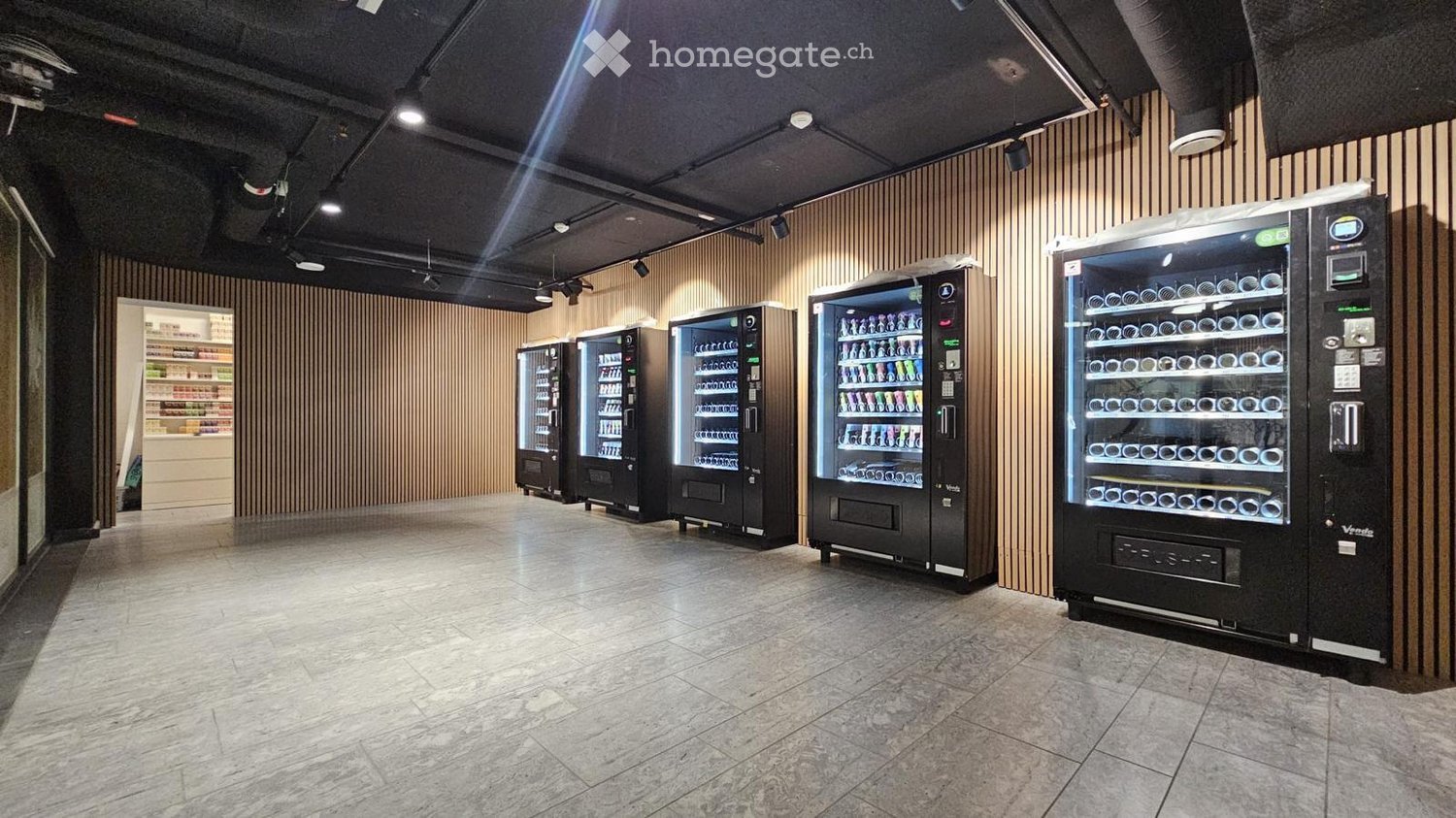 Modern, well-lit room with polished marble flooring and sleek wooden panel walls. Multiple black vending machines are lined up against the wall, each stocked with various beverages and snacks.