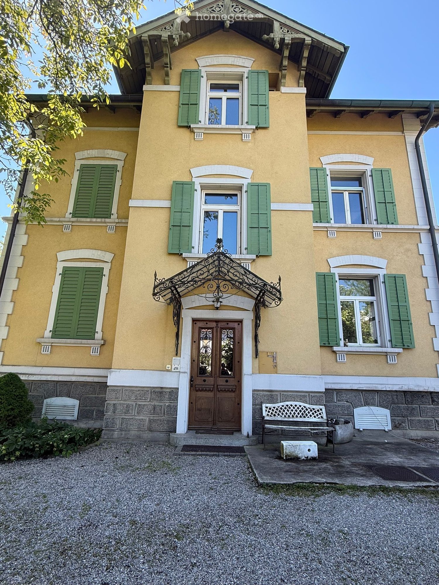 House. Two-story yellow house with green shutters, ornate entrance, stone base, and small garden area with a bench.